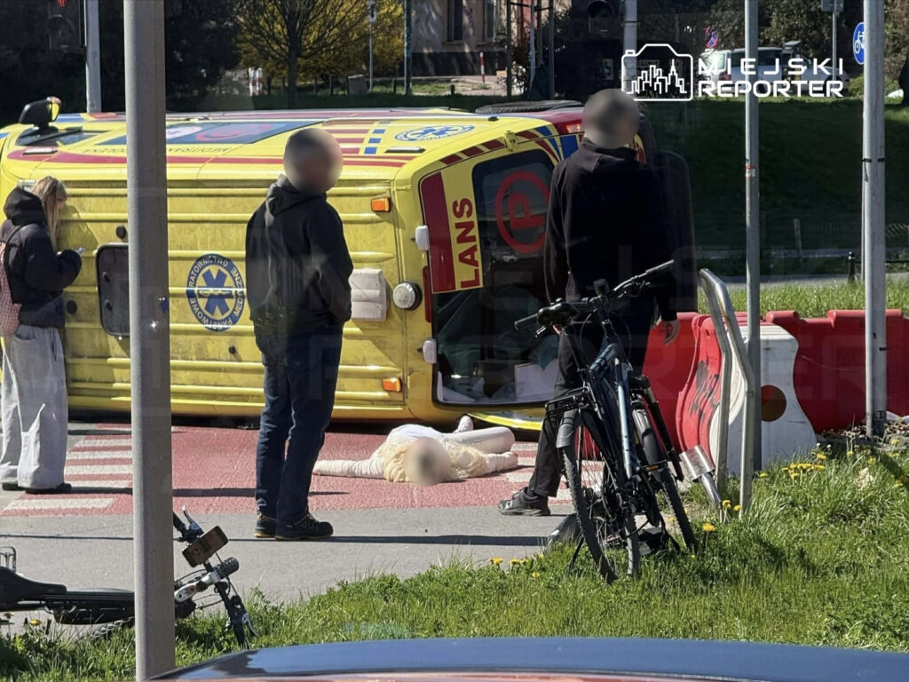 Grupa osób stoi obok przewróconego pojazdu ratunkowego na ulicy, a na jezdni leży osoba.