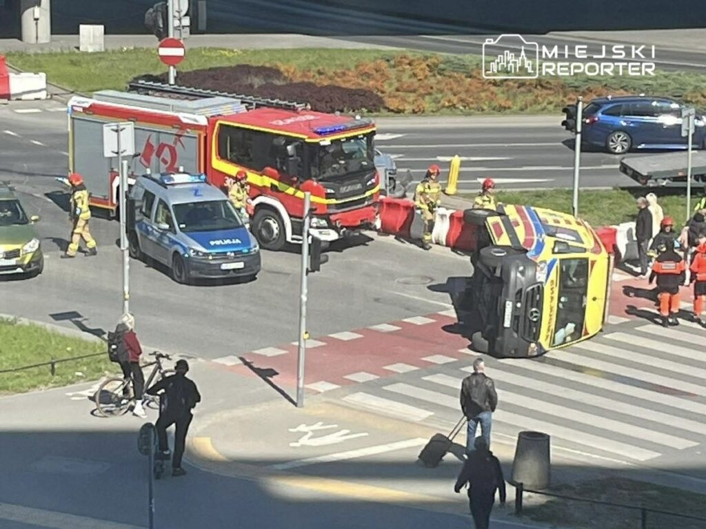 Na skrzyżowaniu stoi przewrócony pojazd, wokół niego interweniują strażacy i policjanci, a przechodnie obserwują sytuację.