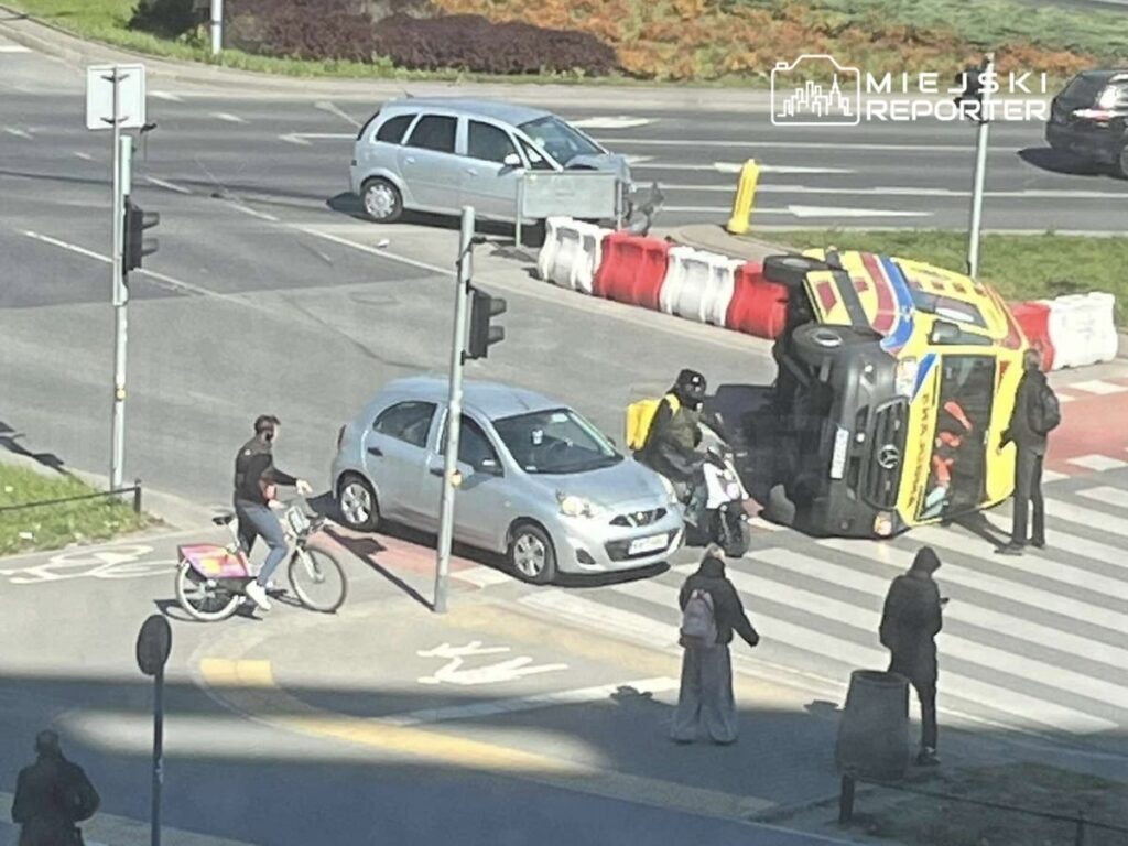 Na przejściu dla pieszych stoi mężczyzna na rowerze, obok przewrócony samochód dostawczy, a w tle inne pojazdy.