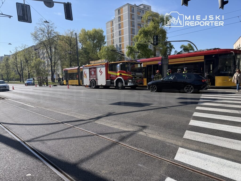 W miejscu zdarzenia stoi wóz strażacki oraz tramwaj, obok grupka strażaków i policjantów. Na jezdni widoczne są pachołki.