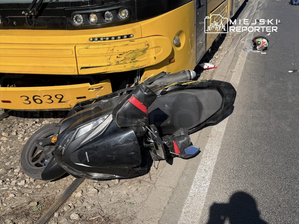 Skuter leży na boku obok żółtego tramwaju, który ma zarysowania na przodzie, na torach w miejskim otoczeniu.