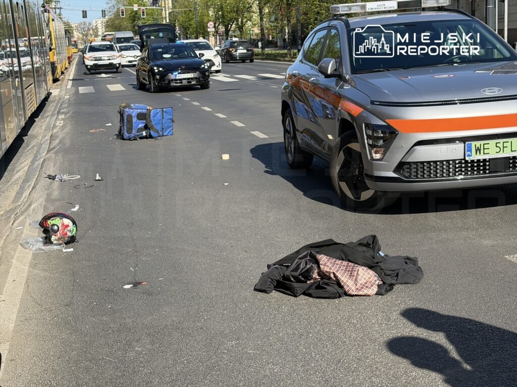 Na jezdni leżą porozrzucane przedmioty, w tym niebieska torba, kask motocyklowy oraz ubrania, obok zatrzymane pojazdy.