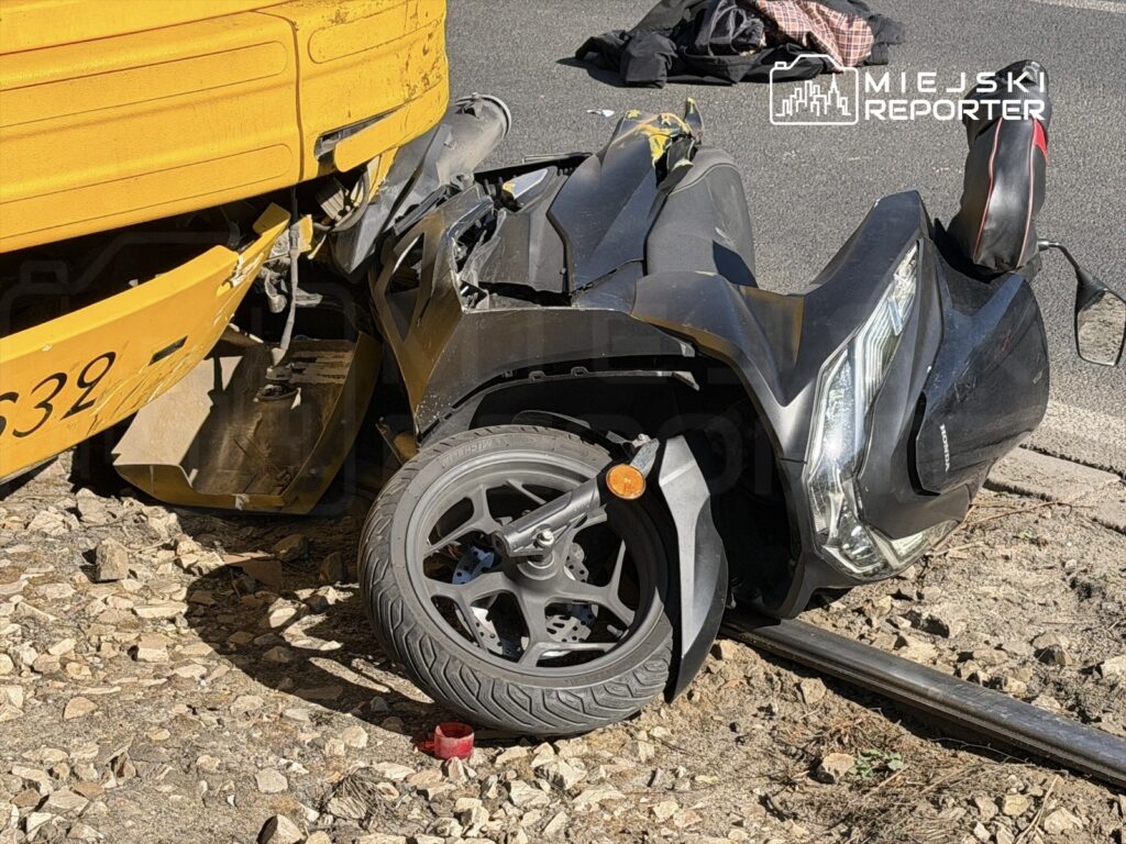 Zniszczony skuter leży obok żółtej ciężarówki na poboczu drogi, wokół leżą kamienie.