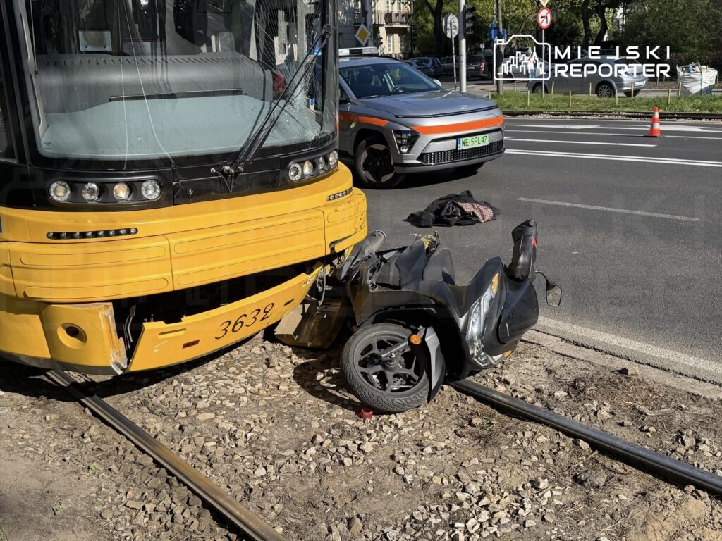Żółty tramwaj stoi na torach, a obok leży uszkodzony skuter. Na drodze widoczne są inne pojazdy oraz oznakowanie drogowe.