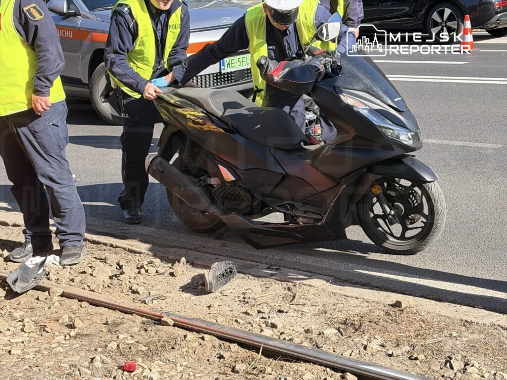 Dwaj policjanci w żółtych kamizelkach pomagają w podnoszeniu czarnego skutera leżącego na poboczu drogi.