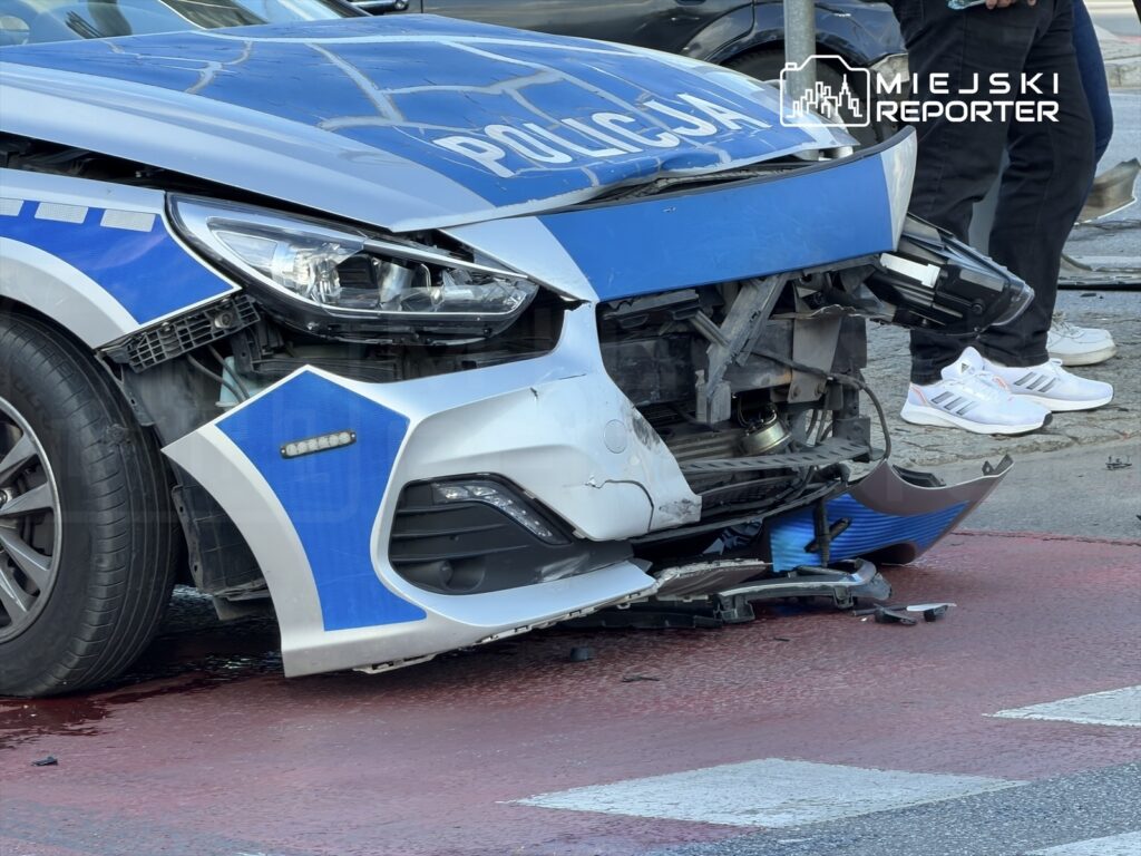 Uszkodzony radiowóz policyjny z uszkodzoną przednią częścią na ulicy, obok stojących osób w białych butach.