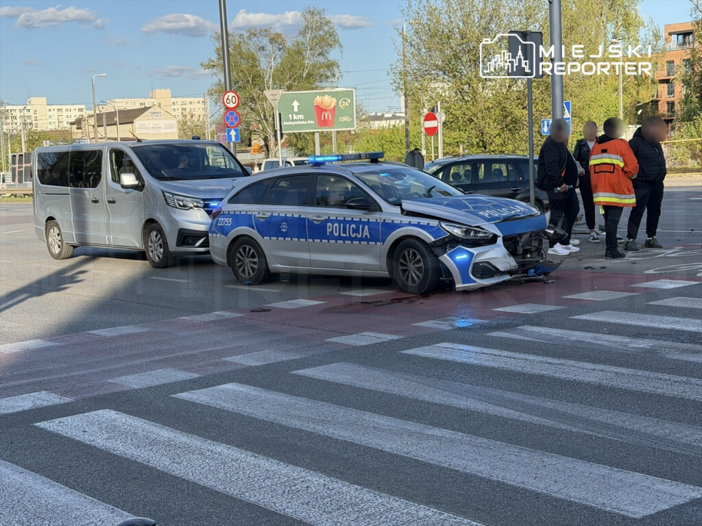 Policyjny radiowóz z uszkodzoną przednią częścią stoi na skrzyżowaniu, obok zatrzymany szary van. Kilku mężczyzn rozmawia w pobliżu.