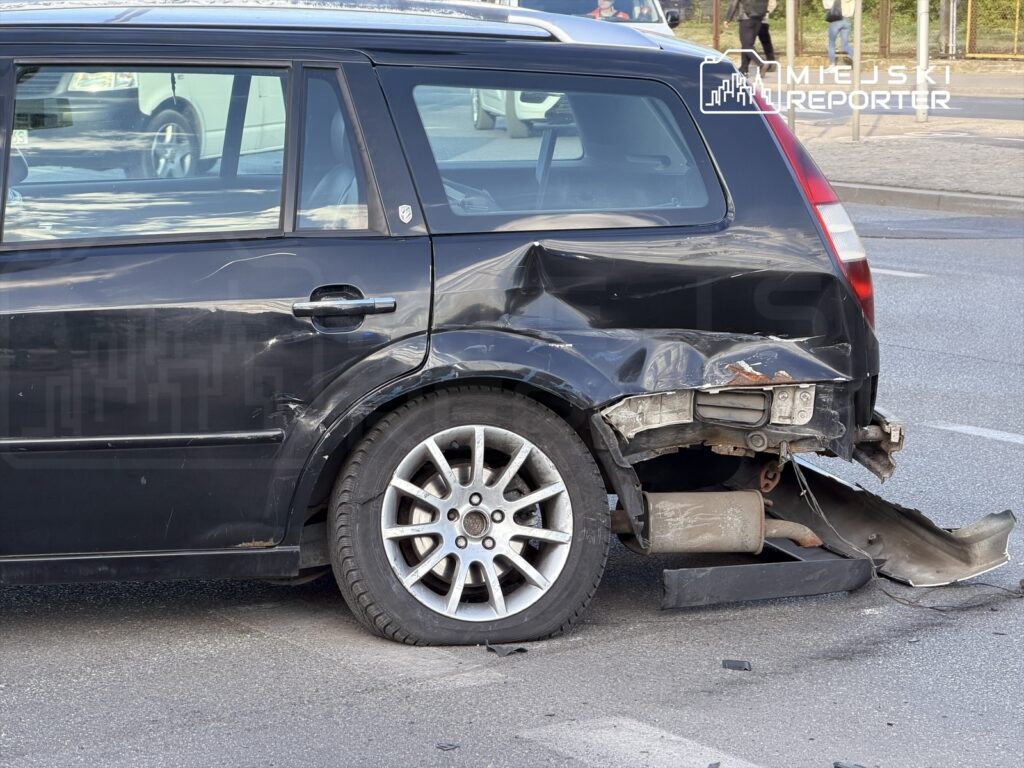 Czarny samochód osobowy z uszkodzoną tylną częścią stoi na ulicy, obok widać inne pojazdy.