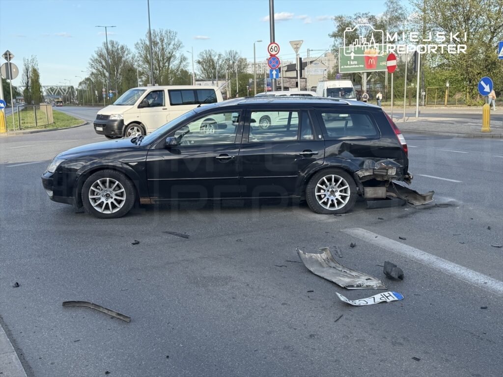 Czarny samochód osobowy z uszkodzoną tylną częścią stoi na skrzyżowaniu, obok leżą fragmenty karoserii.