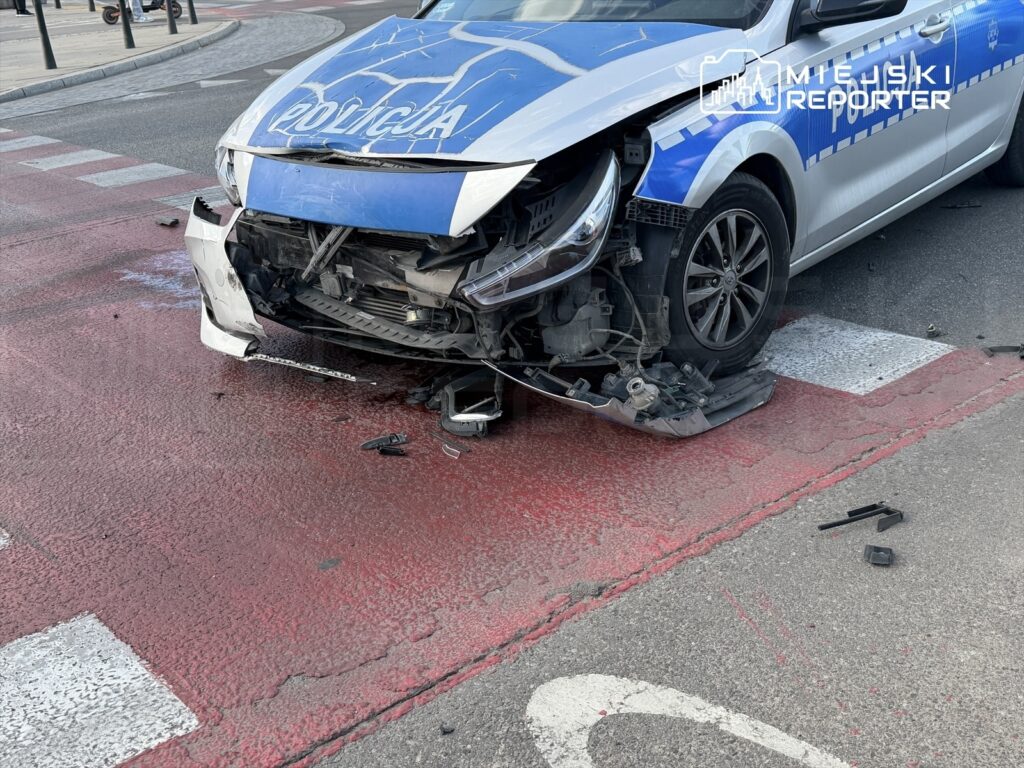 Uszkodzony radiowóz policji z uszkodzoną przednią częścią, zaparkowany na czerwonym pasie drogi.