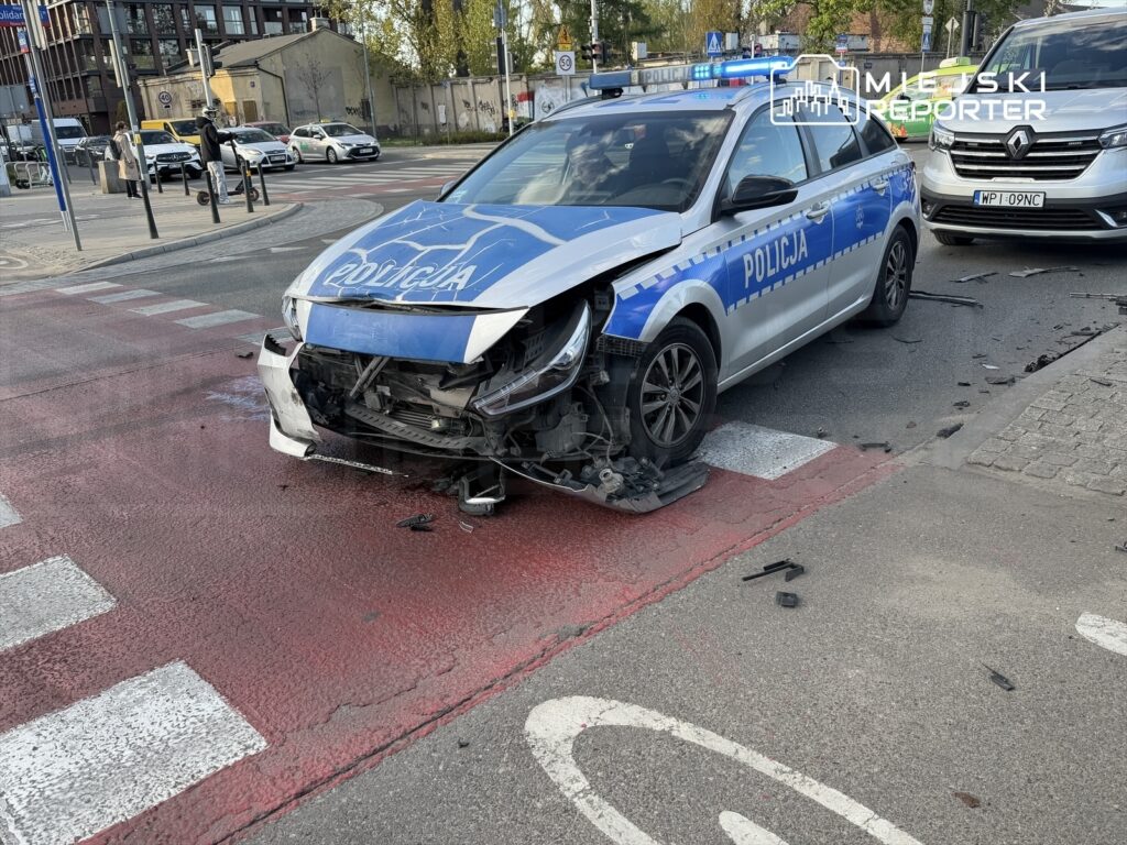 Policyjny samochód z uszkodzoną przednią częścią stoi na czerwonym pasie na ulicy, obok innych pojazdów.