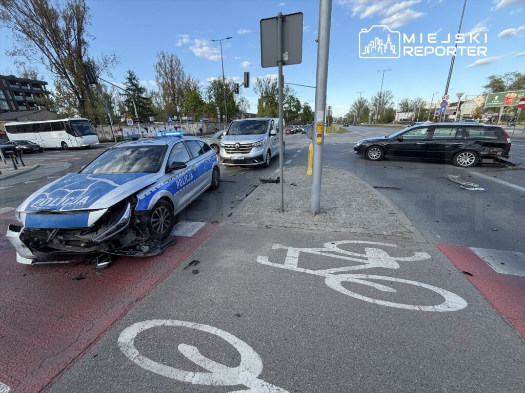 Policyjny radiowóz z uszkodzoną przednią częścią stoi na skrzyżowaniu obok dwóch innych pojazdów po kolizji.