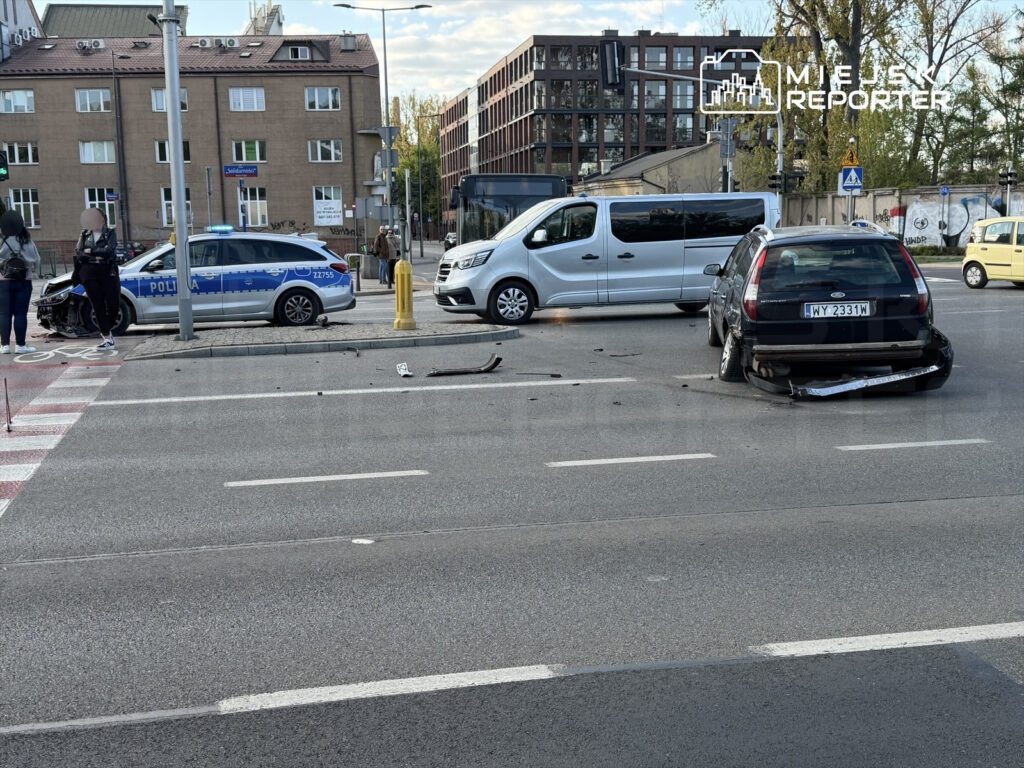 Zderzenie dwóch samochodów na skrzyżowaniu, obok radiowozu policji i pojazdu dostawczego. Na jezdni leżą fragmenty pojazdów.