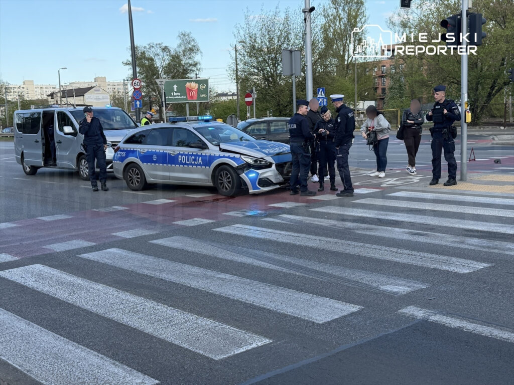 Grupa policjantów prowadzi interwencję na przejściu dla pieszych obok radiowozu z włączonymi sygnałami świetlnymi.