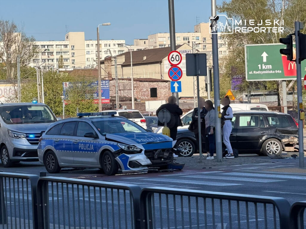 Policyjna radiowóz z uszkodzoną przednią częścią stoi na skrzyżowaniu, obok grupy ludzi rozmawiających przy znaku drogowym.