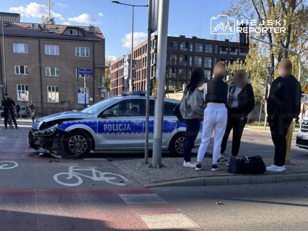 Policjant rozmawia z grupą osób obok uszkodzonego radiowozu na skrzyżowaniu w mieście.