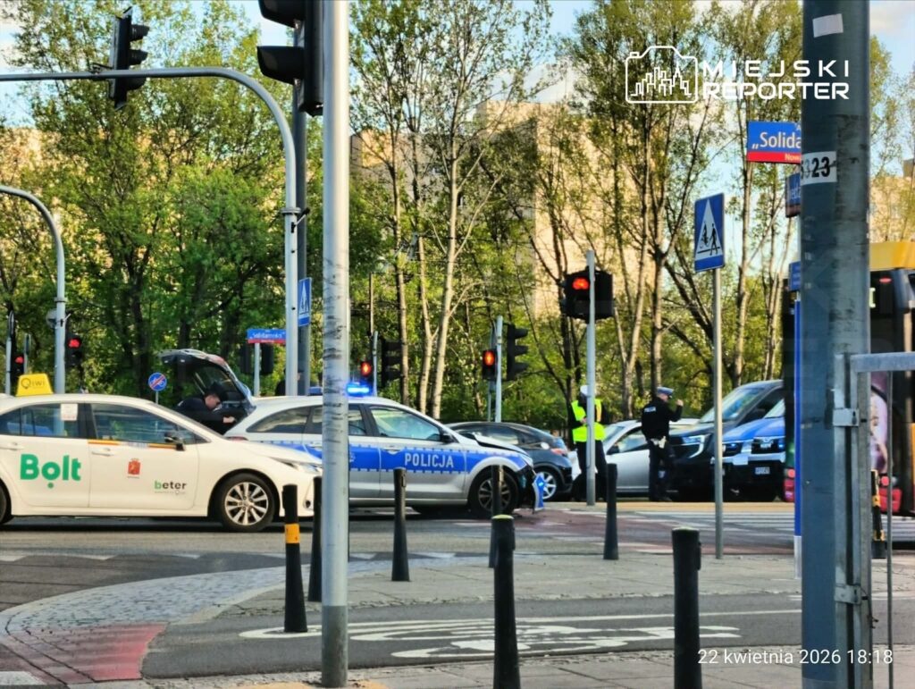 Policyjny radiowóz z włączonymi sygnalizatorami stoi na skrzyżowaniu, a policjant rozmawia z kierowcą taksówki.