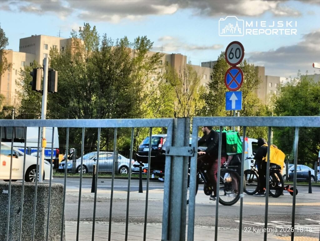 Rowerzyści przejeżdżają obok znaku ograniczenia prędkości do 60 km/h, w tle nowoczesne budynki i zieleń.