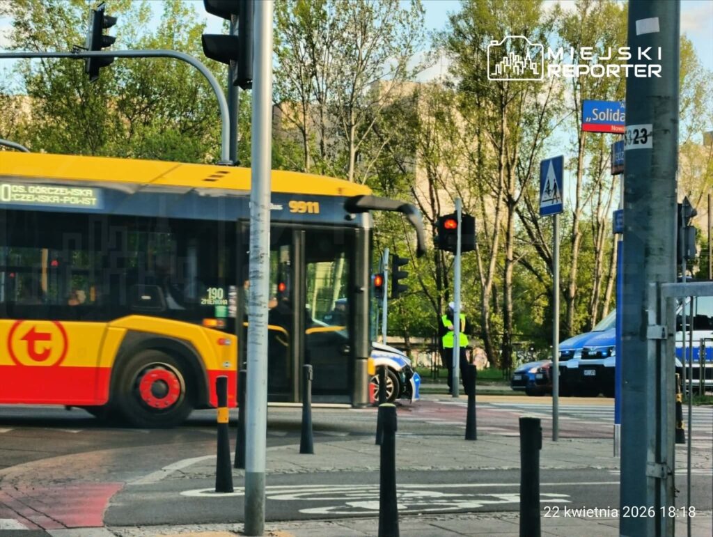 Żółty autobus komunikacji miejskiej skręca w prawo na skrzyżowaniu, obok strażnika w kamizelce odblaskowej.