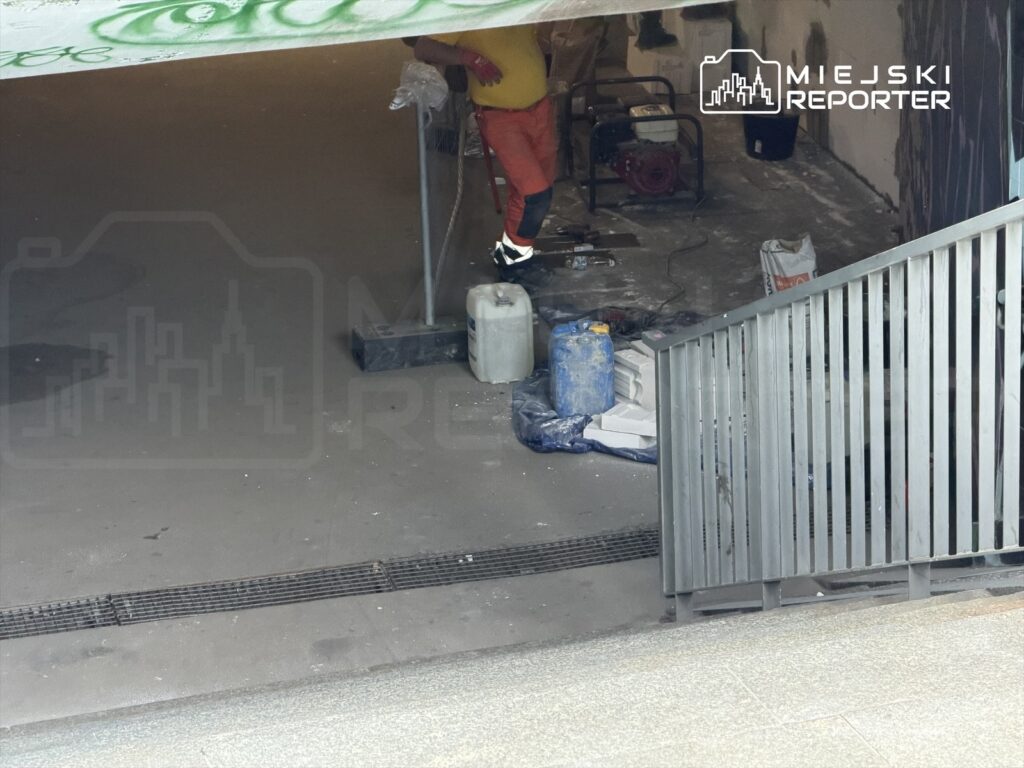 Mężczyzna w żółtej koszulce maluje ścianę w podziemnym pomieszczeniu, otoczony narzędziami i materiałami budowlanymi.