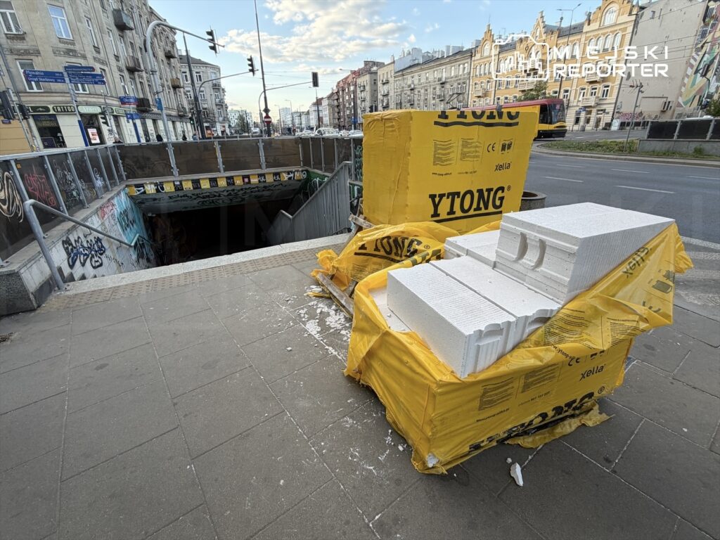 Na chodniku obok wejścia do metra leżą opakowania materiałów budowlanych, w tym białe bloczki i żółte torby.