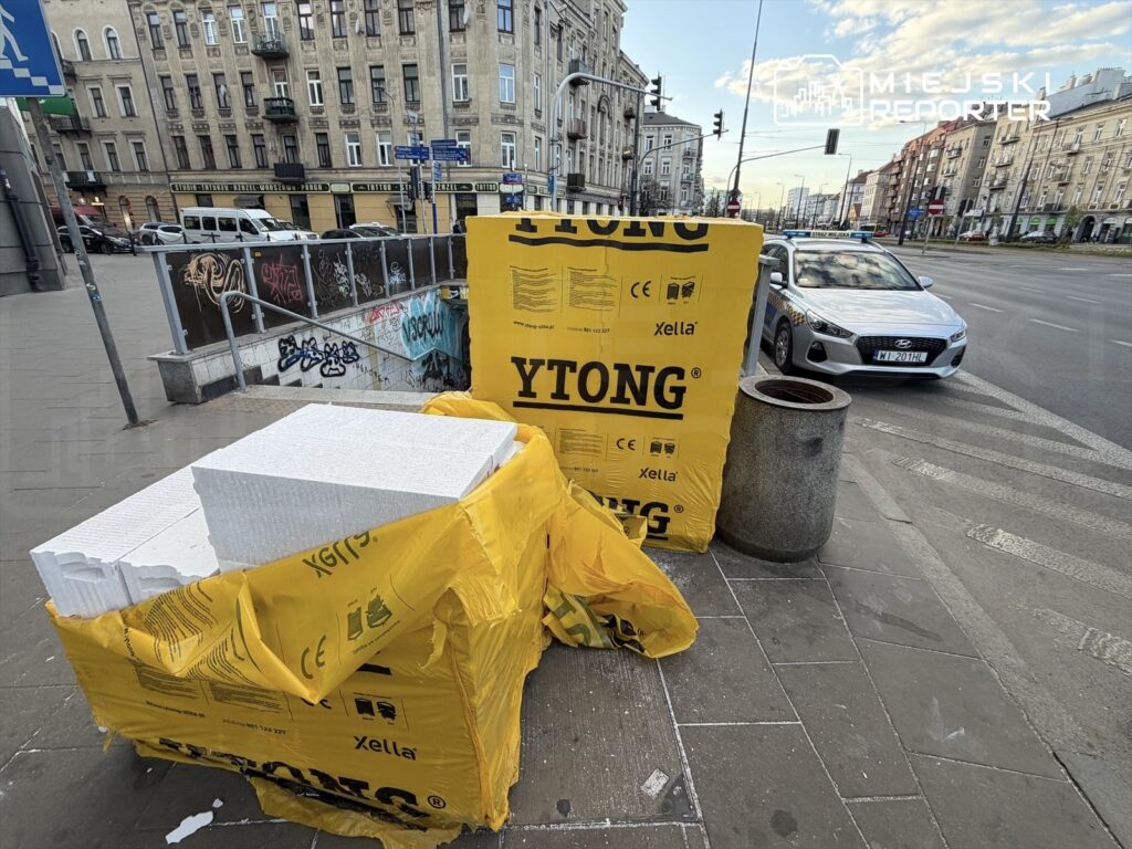 Na chodniku leżą kartony z białymi bloczkami styropianowymi, obok stoi kosz na śmieci, w tle widoczna ulica.