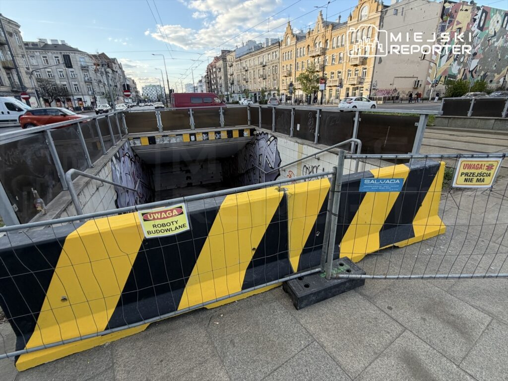 Otwór wykopany w jezdni, ogrodzony barierkami z oznaczeniami „UWAGA ROBOTY BUDOWLANE” i „UWAGA! PRZEJŚCIA NIE MA”. W tle widoczne kamienice.
