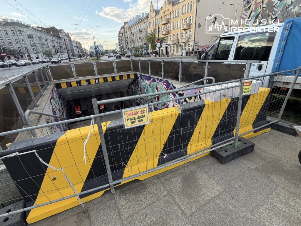 Robotnicy w odblaskowych kamizelkach pracują przy wejściu do wykopu, otoczonym barierkami i znakami ostrzegawczymi.