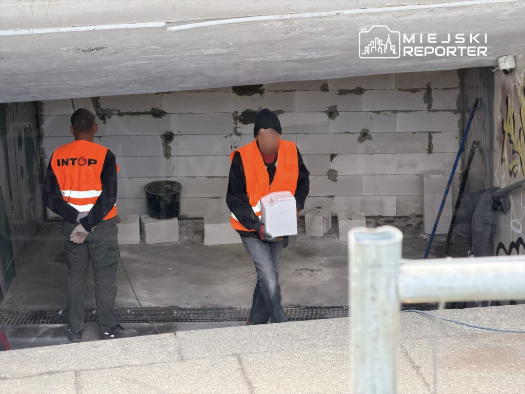 Dwaj mężczyźni w pomarańczowych kamizelkach roboczych przenoszą materiały budowlane w nieukończonym pomieszczeniu.