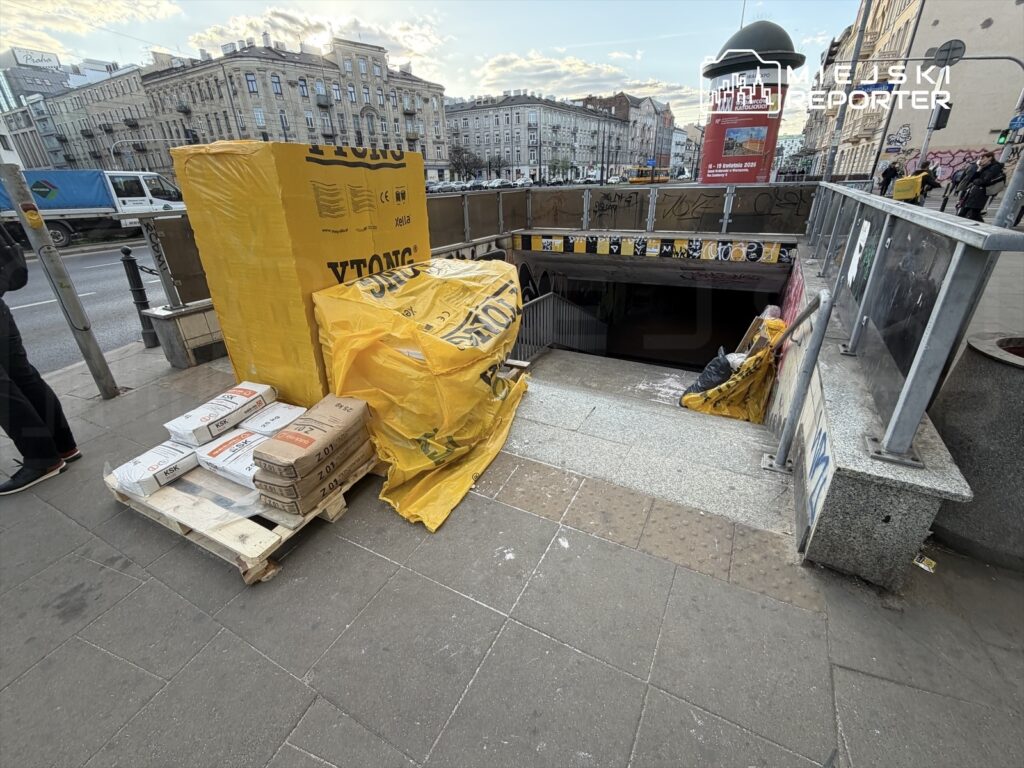 Paczki materiałów budowlanych, w tym bloczki Ytong, leżą na palecie obok wejścia do stacji metra, otoczone folią.