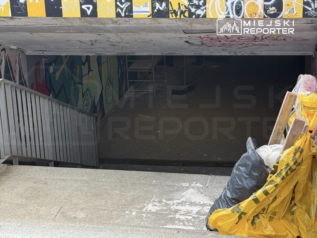 Schody prowadzące do ciemnego przejścia z graffiti na ścianach, z workami na śmieci po prawej stronie.