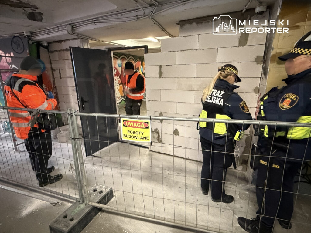 Pracownicy budowlani oraz straż miejska kontrolują teren robót budowlanych, wokół ogrodzenia z siatki.