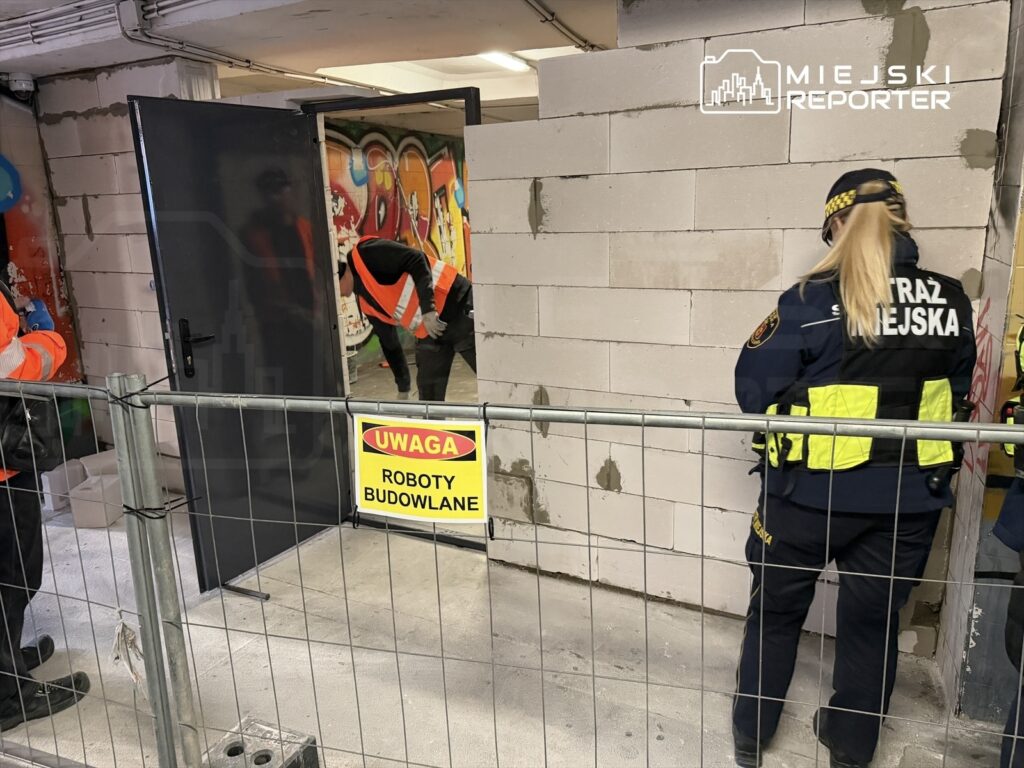 Pracownicy budowlani w kamizelkach odblaskowych prowadzą prace w pomieszczeniu z graffiti, a obok stoi straż miejska.