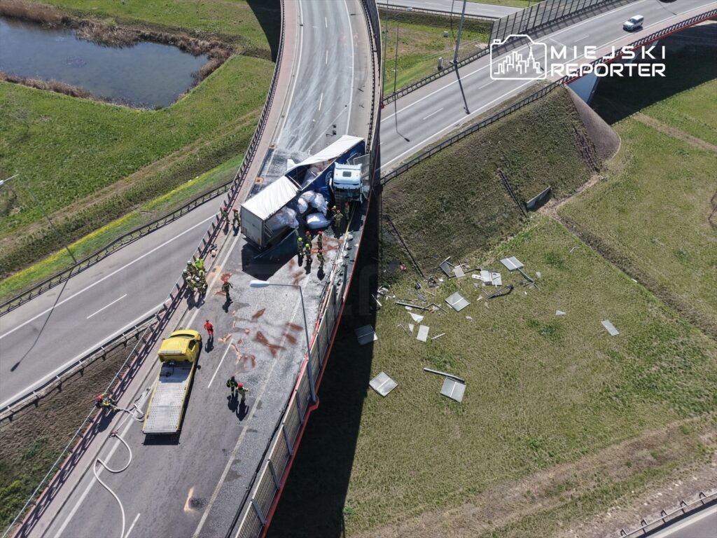 Wielu ratowników pracuje przy przewróconej ciężarówce na skrzyżowaniu dróg, obok leżących uszkodzonych znaków drogowych.