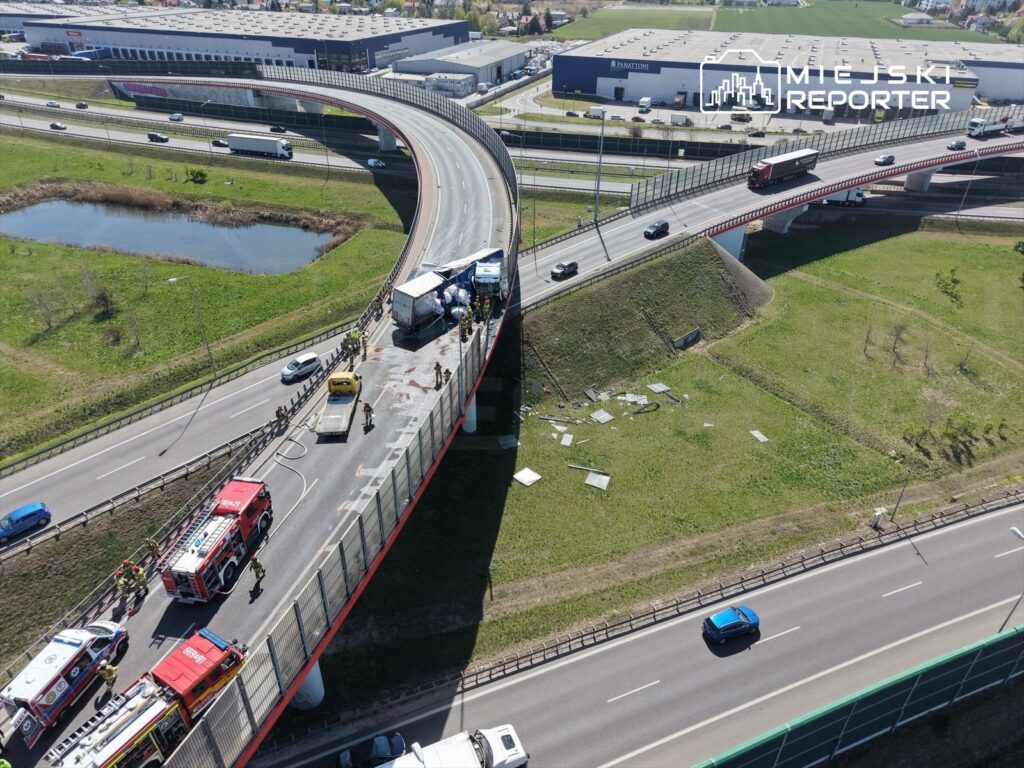 Wielu ratowników pracuje przy wypadku drogowym na estakadzie, obok przewróconej ciężarówki z towarem.