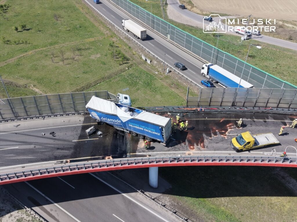 Wielka ciężarówka przewożąca kontener leży na boku na drodze, a ratownicy pracują nad usunięciem przeszkody.
