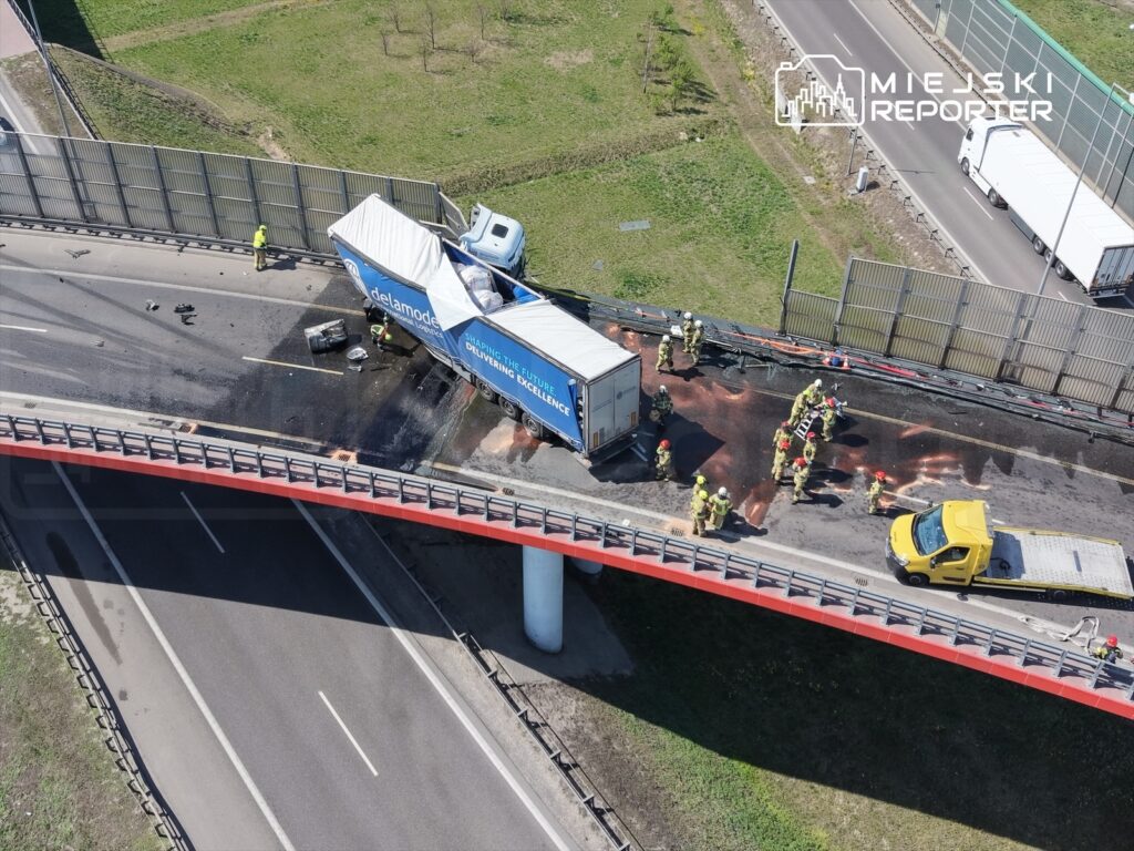 Wielu strażaków pracuje przy wypadku drogowym na moście, gdzie przewróciła się ciężarówka, obok stoi żółty pojazd.