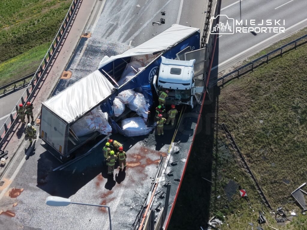 Strażacy pracują nad wypadkiem drogowym z ciężarówką, która przewróciła się na jezdni, obok uszkodzonego barierki ochronnej.
