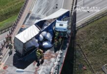 Strażacy pracują nad wypadkiem drogowym z ciężarówką, która przewróciła się na jezdni, obok uszkodzonego barierki ochronnej.