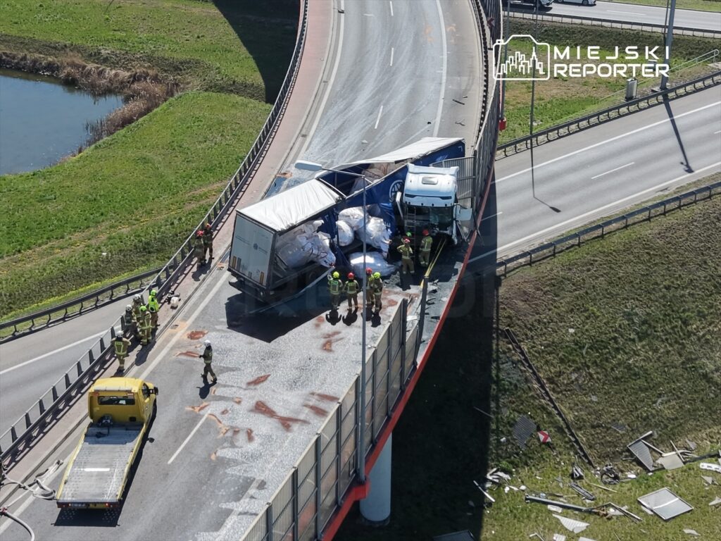 Ciężarówka przewożąca materiały budowlane przewrócona na zakręcie, a obok pracują strażacy i służby ratunkowe.