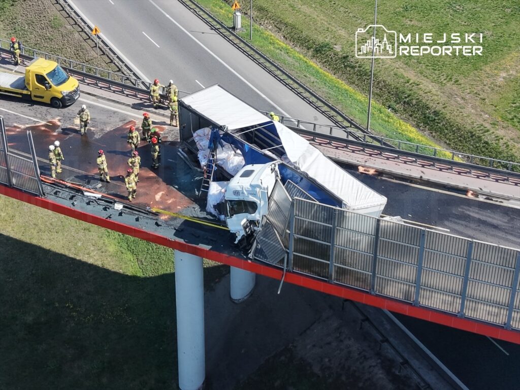 Grupa strażaków zabezpiecza miejsce wypadku na moście, gdzie ciężarówka przewróciła się, a załadunek jest rozrzucony na jezdni.