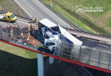 Grupa strażaków zabezpiecza miejsce wypadku na moście, gdzie ciężarówka przewróciła się, a załadunek jest rozrzucony na jezdni.