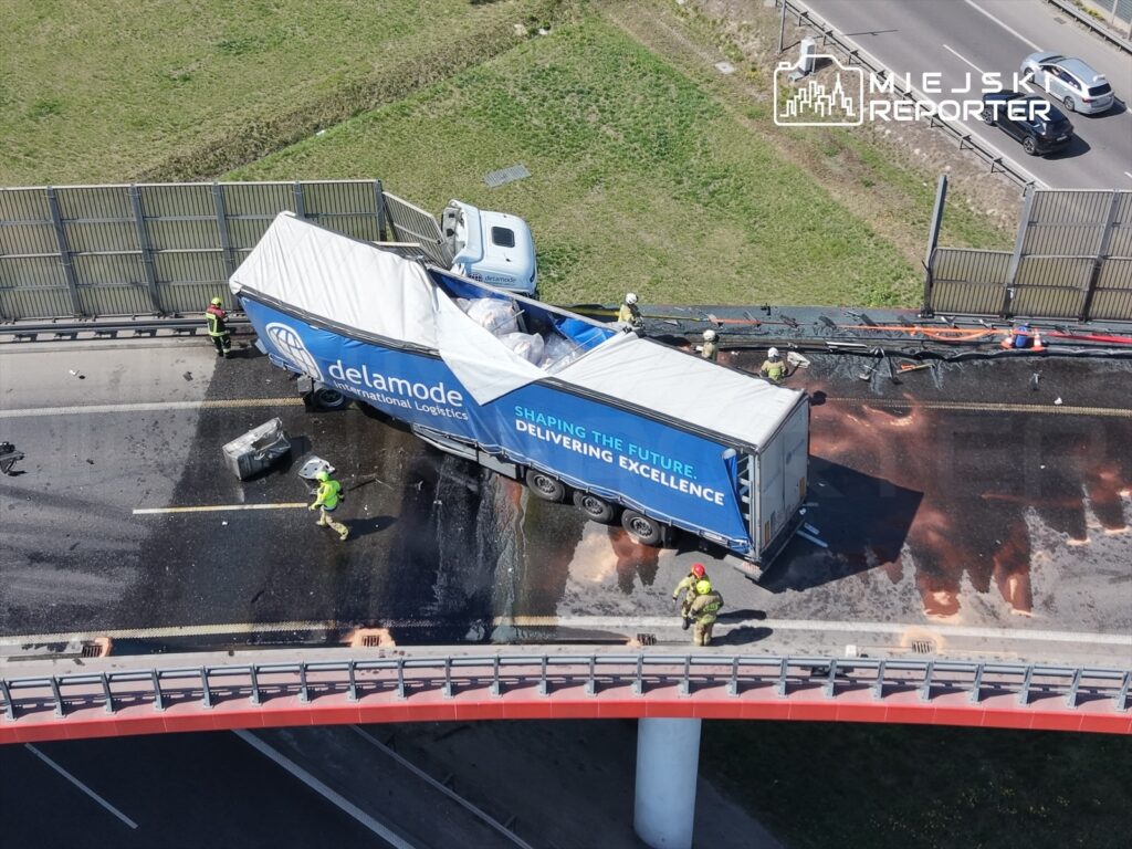 Ciężarówka z logo "delamode" przewrócona na jezdni, obok pracują strażacy w kaskach i odblaskowych kamizelkach.