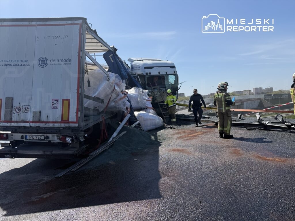 Wypadek drogowy z udziałem ciężarówki, która przewróciła się na jezdni, obok pracujący ratownicy i policjanci.