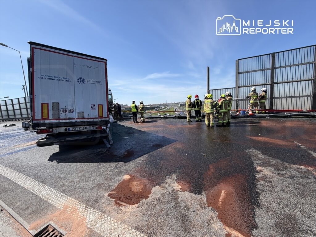 Zespół strażaków pracuje nad usunięciem plamy substancji na drodze, obok ciężarówki na autostradzie.