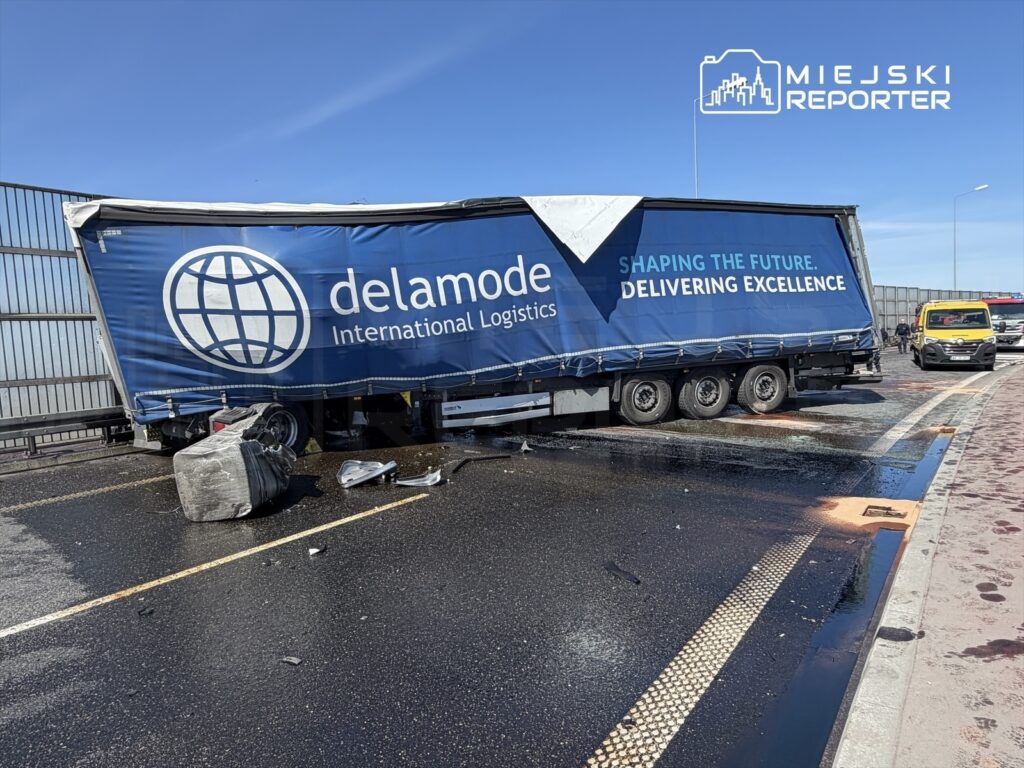 Ciągnik siodłowy z naczepą firmy Delamode, uszkodzony, stojący na drodze, obok rozbite elementy oraz plama oleju.