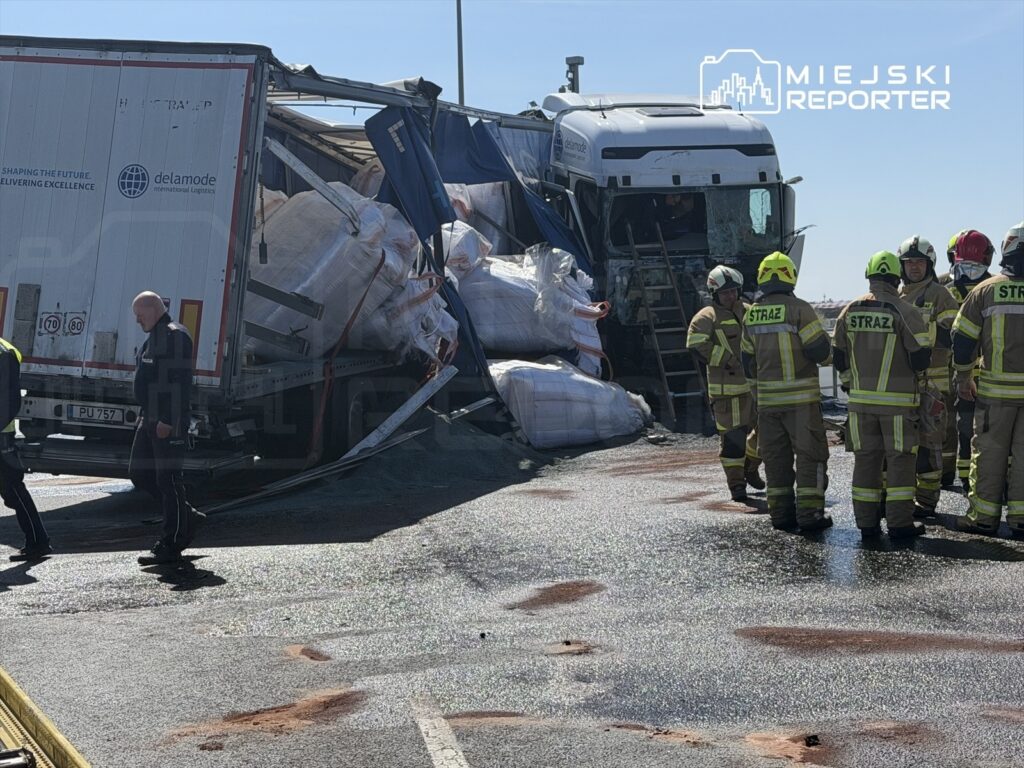 Ciągnik siodłowy z przewróconą naczepą, a obok zespół strażaków omawiających działania ratunkowe na drodze.