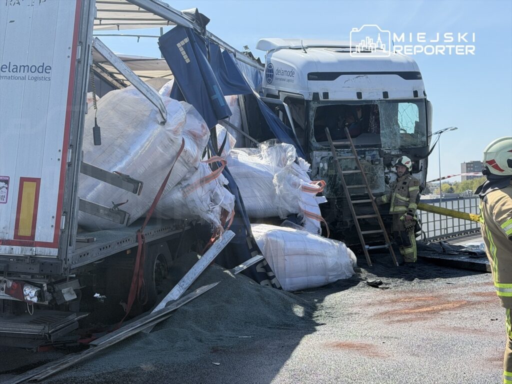 Ciężarówka z uszkodzonym przodem leży na boku, a strażacy zabezpieczają ładunek z workami na drodze.