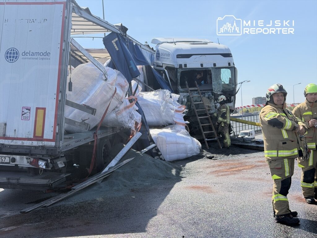 Ciężarówka z uszkodzoną naczepą leży na jezdni, a strażacy zabezpieczają miejsce zdarzenia.