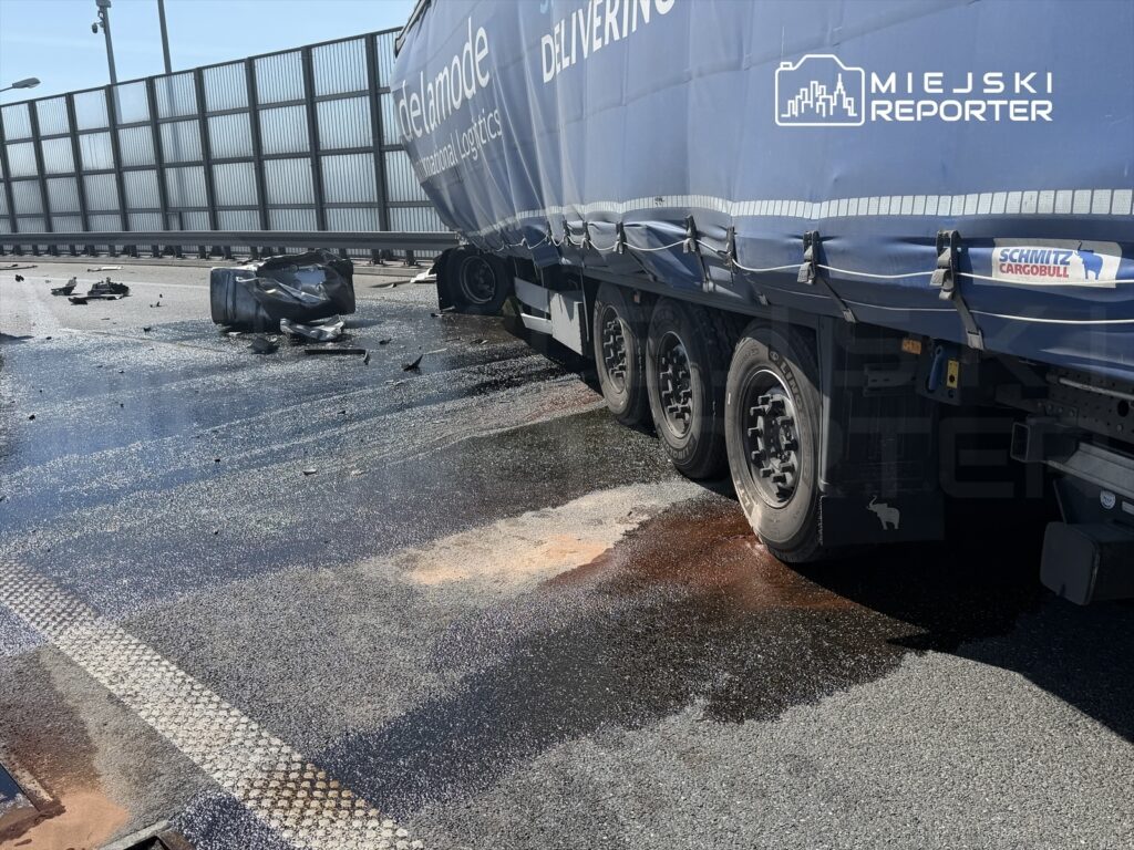 Uszkodzony kontener leży na drodze obok ciężarówki, z rozlanym płynem i odłamkami wokół.